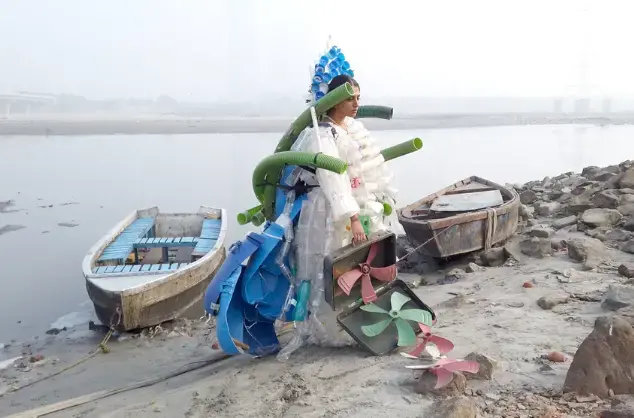 A woman sits on the banks of a wide river surrounded by abandoned boats and rocks. She wears a costume made from plastic bottles, tubing and machinery parts.