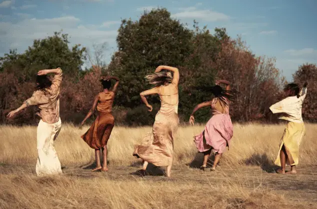 Five women in flowing dresses dance outside in nature