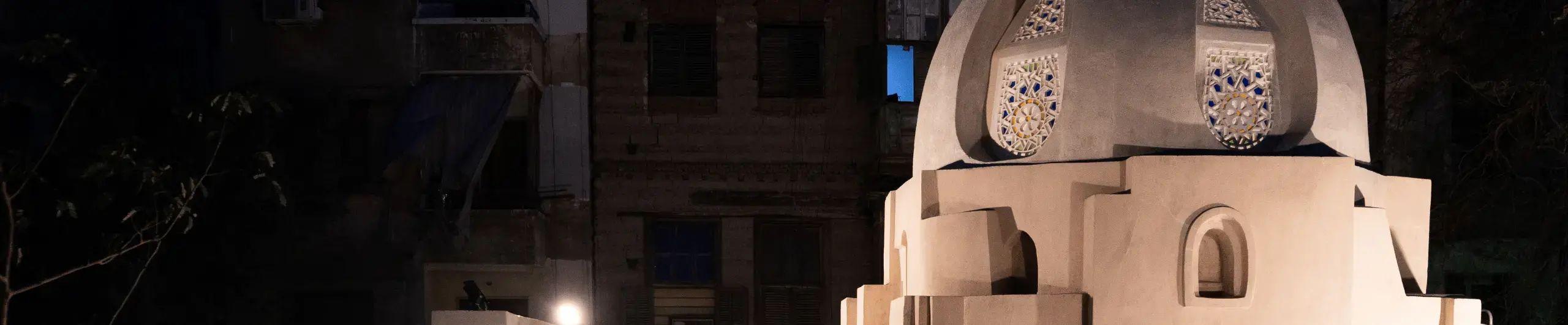 Photograph of Safiyy al-Din Jawhar, a restored medieval Islamic monument in Cairo taken at night. The focus is on the courtyard which has plants.