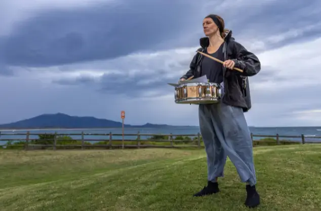 A woman plays a snare drum, hung around her neck. She is standing on a grassy mound looking outwards to the left of the image, and behind her is a body of water with a hill in the distance under a moody cloudy sky