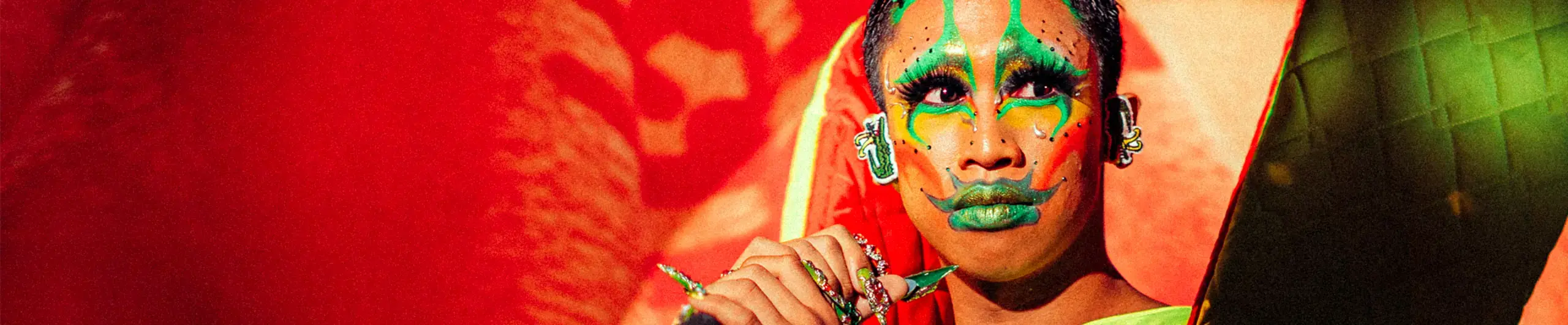 A performer in Filipino traditional dress and bright green face paints stands on stage holding a microphone on a stand, and looks out toward the right of the image