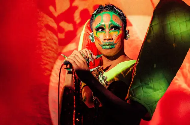 A performer in Filipino traditional dress and bright green face paints stands on stage holding a microphone on a stand, and looks out toward the right of the image