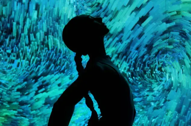 The silhouette of a dancer poses with their head and neck arched backwards and arms outstretched behind them, with long hair tied in a plait. The figure stands in front of an illuminated backdrop with swirling colours of blues, greens and turquoise.
