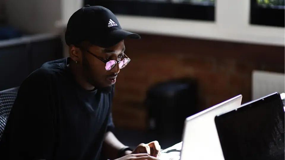 Young man wearing a black Adidas cap and glasses working on a laptop indoors.