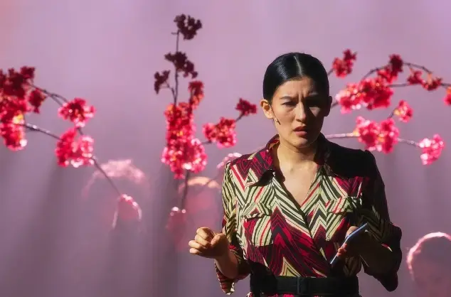 A production image from Confessions of a Shinagawa Monkey showing a Japanese woman stands on stage in front of a cherry blossom tree and purple stage lights create a mist behind her.