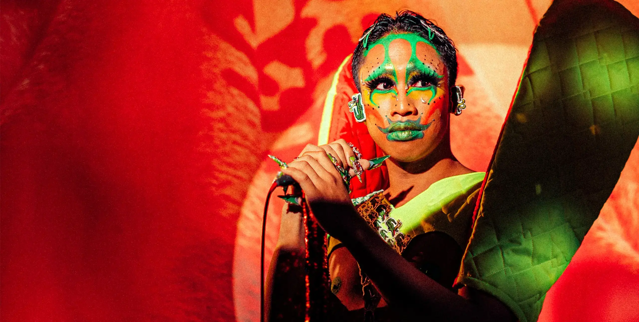A performer in Filipino traditional dress and bright green face paints stands on stage holding a microphone on a stand, and looks out toward the right of the image