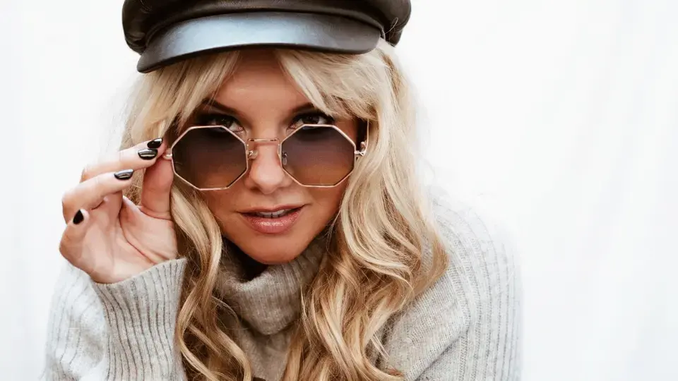 Blonde woman with wavy hair wearing a black leather cap and octagonal sunglasses, adjusting the glasses with her hand.