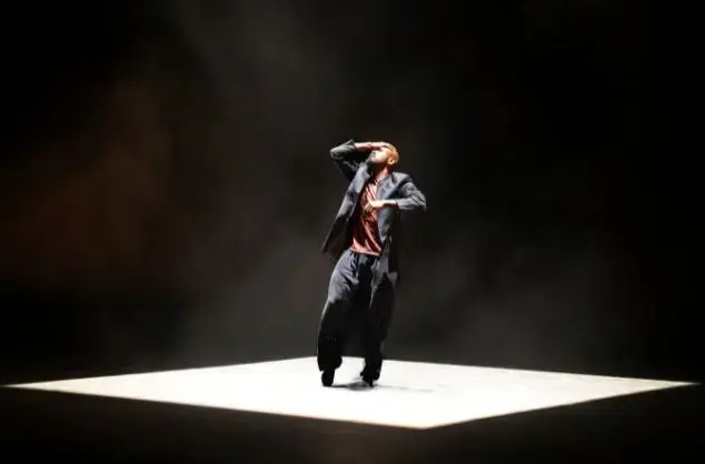 Dickson Mbi performs a dance work standing on a spotlit stage