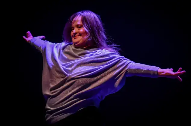 A female performer stands with her arms oustretched, smiling. She is wearing a silver coloured top and is smiling.