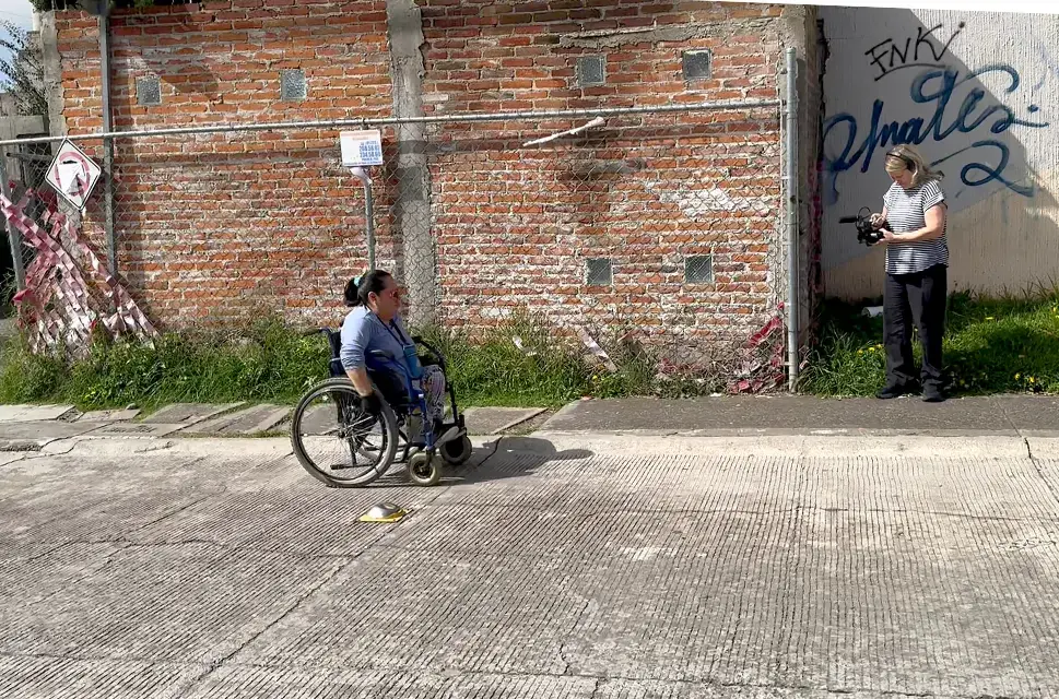 Lady in wheelchair moving from left to right of the picture. On the right stands a lady with a video camera. Behind her is graffiti on a wall.