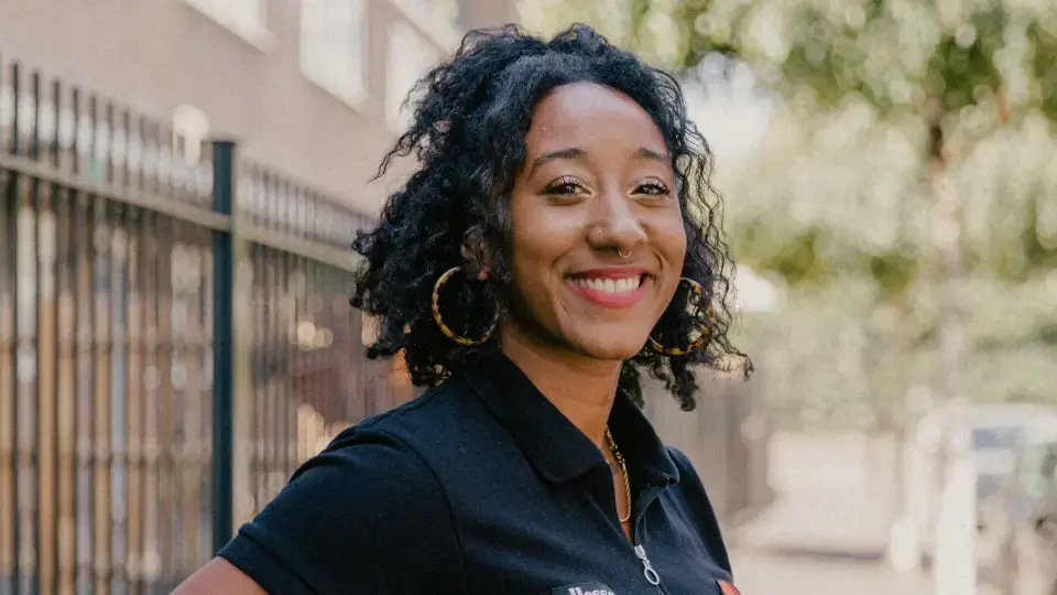 Young woman with curly hair and hoop earrings smiling outdoors near a metal fence and trees.