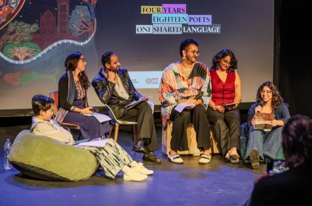 Six people sit on a stage, some are on beanbags, others on low chairs, others on the floor. They are in conversation with each other and are smiling.