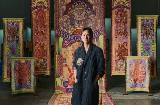 A man stands in front of intricately woven textiles in various colours