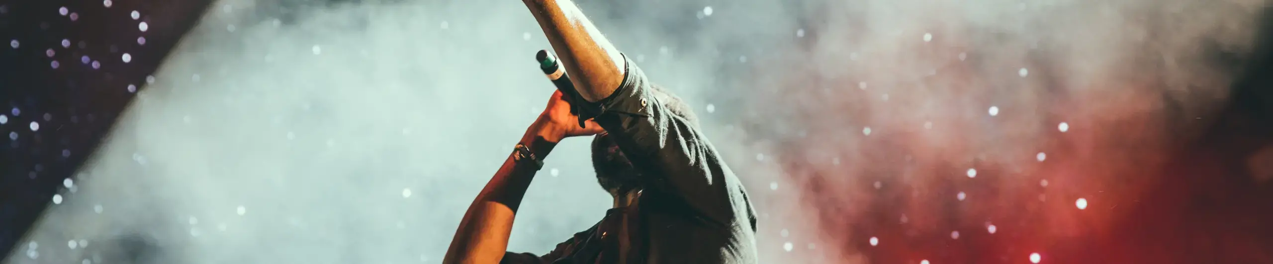 A performer holds a microphone with his hand held up to the sky, in front of a smokey background lit by stage lighting