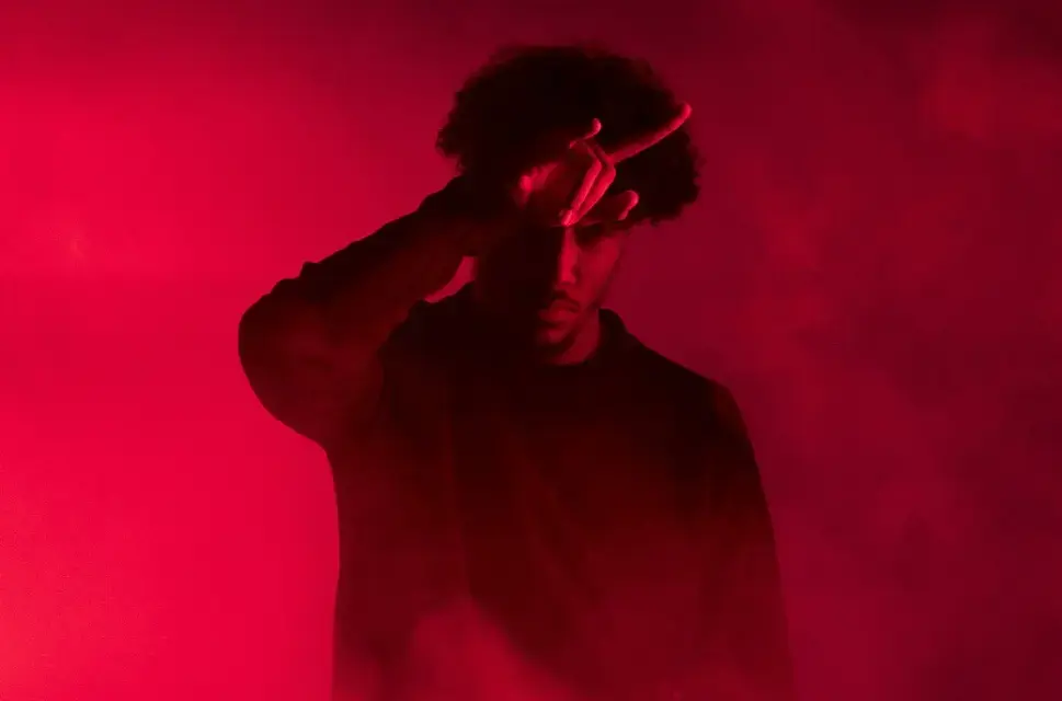 Man in dark red light, holding his right hand against his head.