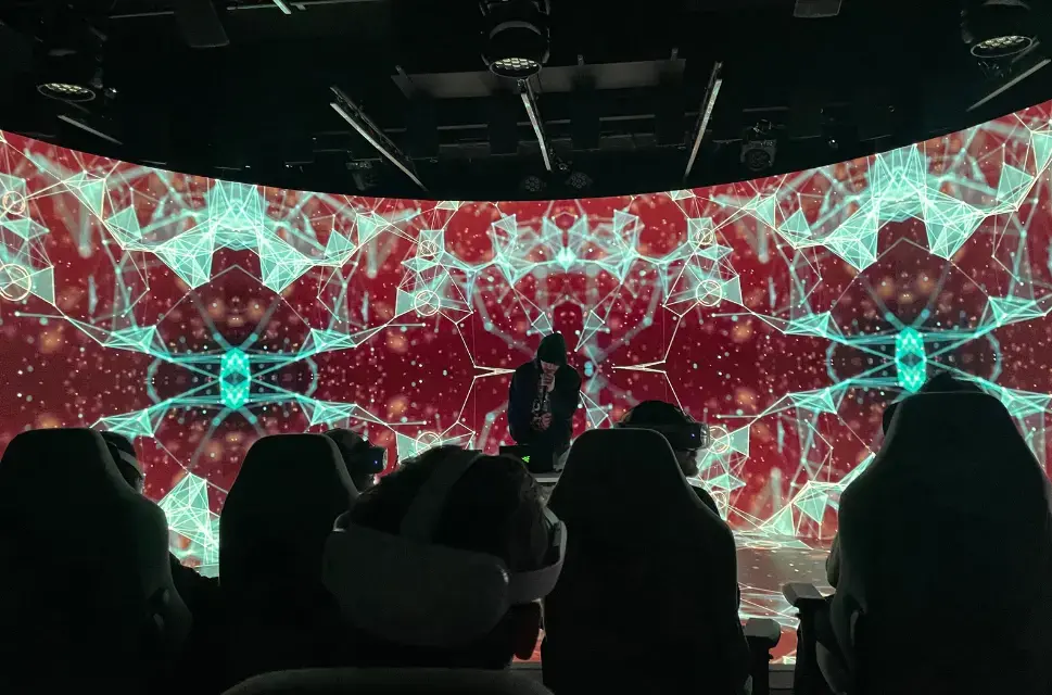 Man on stage holding mic in front of bright lights on the walls behind him. The audience are wearing 3D headsets.