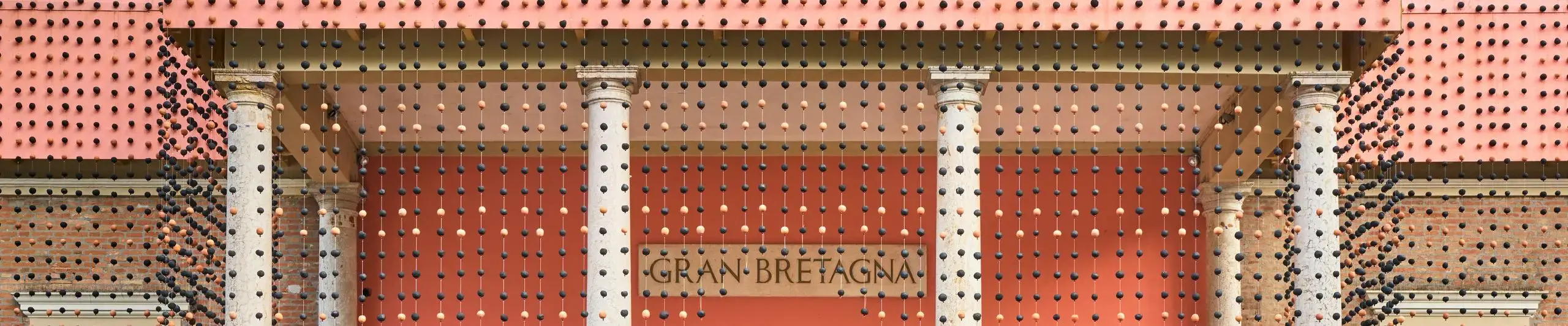 A small section of the view of the front of the British Pavilion at Venice 2025, with a veil of clay beads surrounding the building.