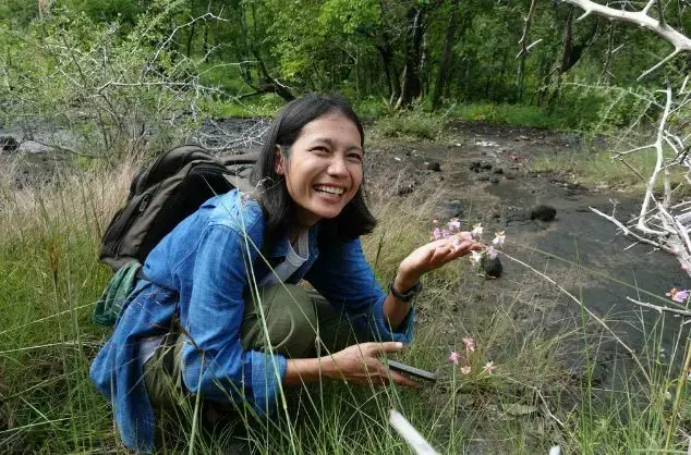 Woman crouches on the ground in long grass in a woodland setting