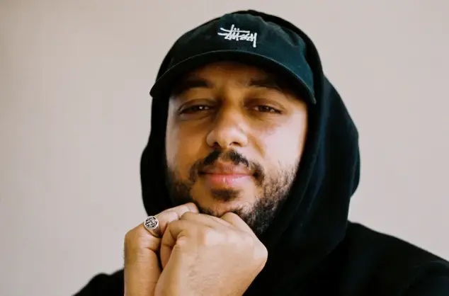Man smiling, wearing a black cap and black hooded jumper, head resting on hands, 