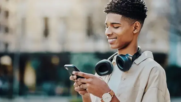 African teenager, smiling, headphones around his neck, typing on his mobile in the city street of Singapore.