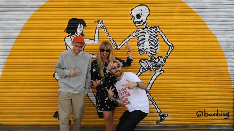 Group of three people smiling and making hand gestures in front of street art on a corrugated metal wall showing a skeleton and a woman dancing.