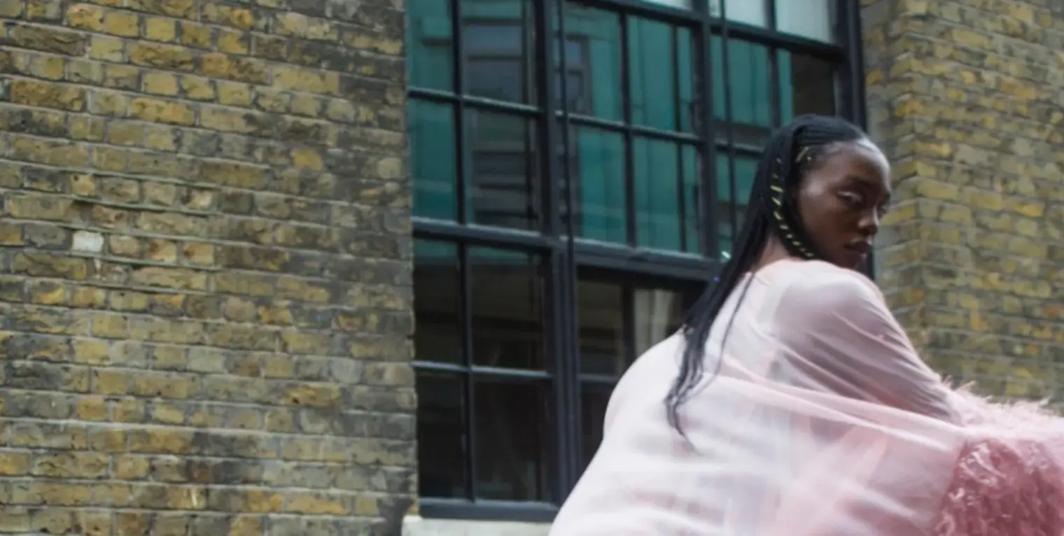 A model walks past a brick building dressed in extravagant pale pink furs and looks back at the camera.