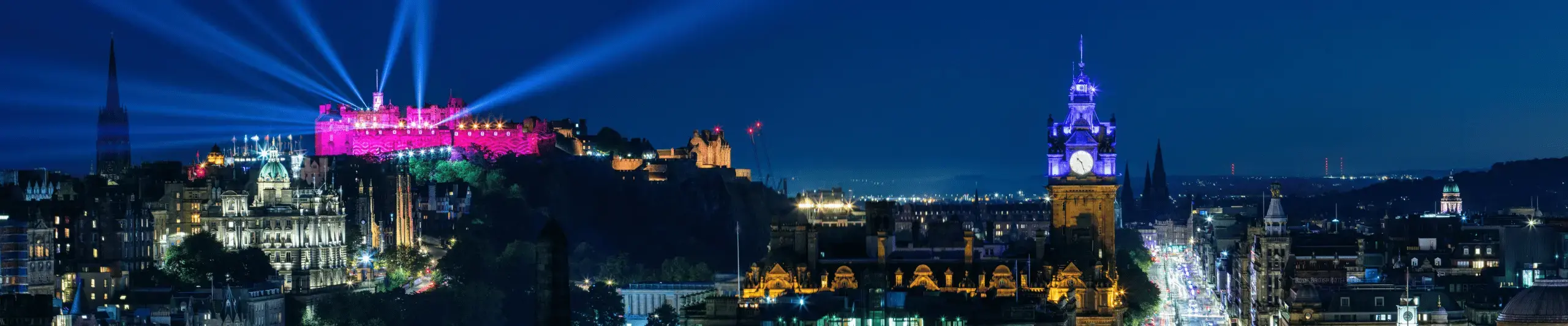 The Edinburgh city skyline at night, taken from Calton Hill, showing Edinburgh Caslte on a hilltop illuminated in pink light with search lights going in all directions. A brightly lit busy street is on the right of the image, with an illuminated clock tower to its left.