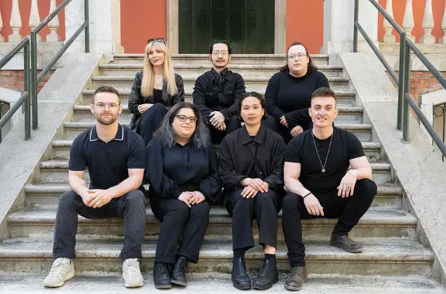 A group of seven people dressed in black sit on the steps of the British Pavilion at the Venice Biennale.