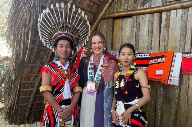 Mari Mathias at Hornbill Festival 2024. Three people, two women and one man, stand together in front of a wooden building. They are dressed in traditional Naga dress.