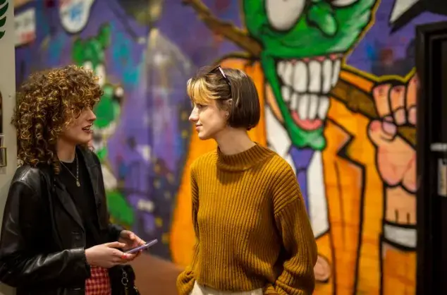 Two young women chatting in front of a wall decorated with bright murals of a cartoonish green face with grinning teeth.