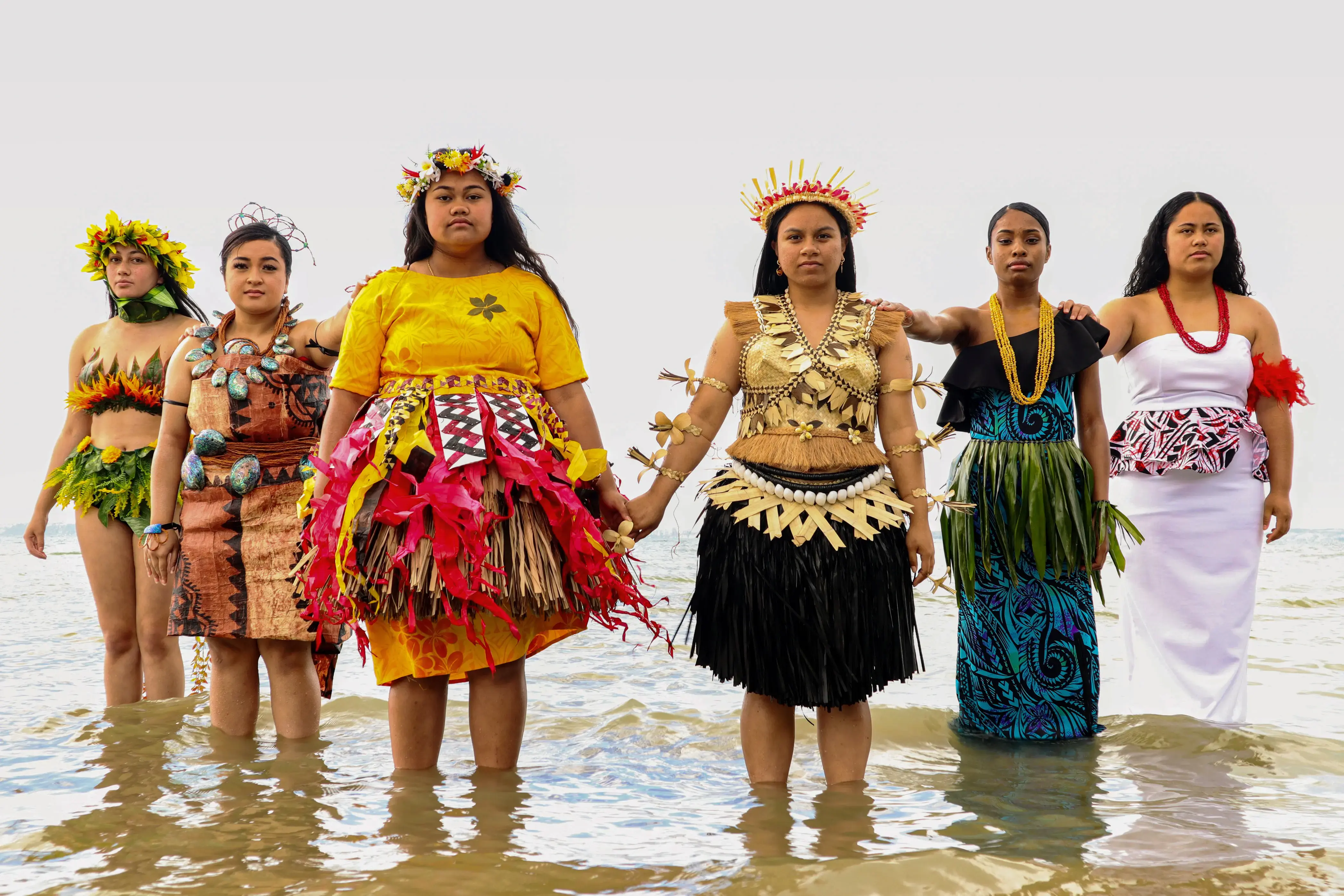 Six young women in colourful traditional dress stand in the sea, hand in hand