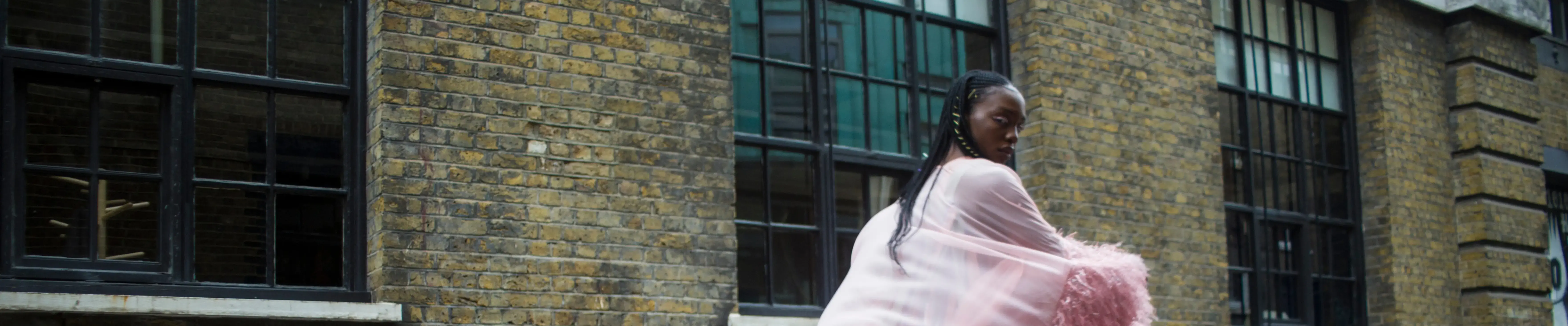 a model walks past a brick building dressed in extravagant pale pink furs and looks back at the camera