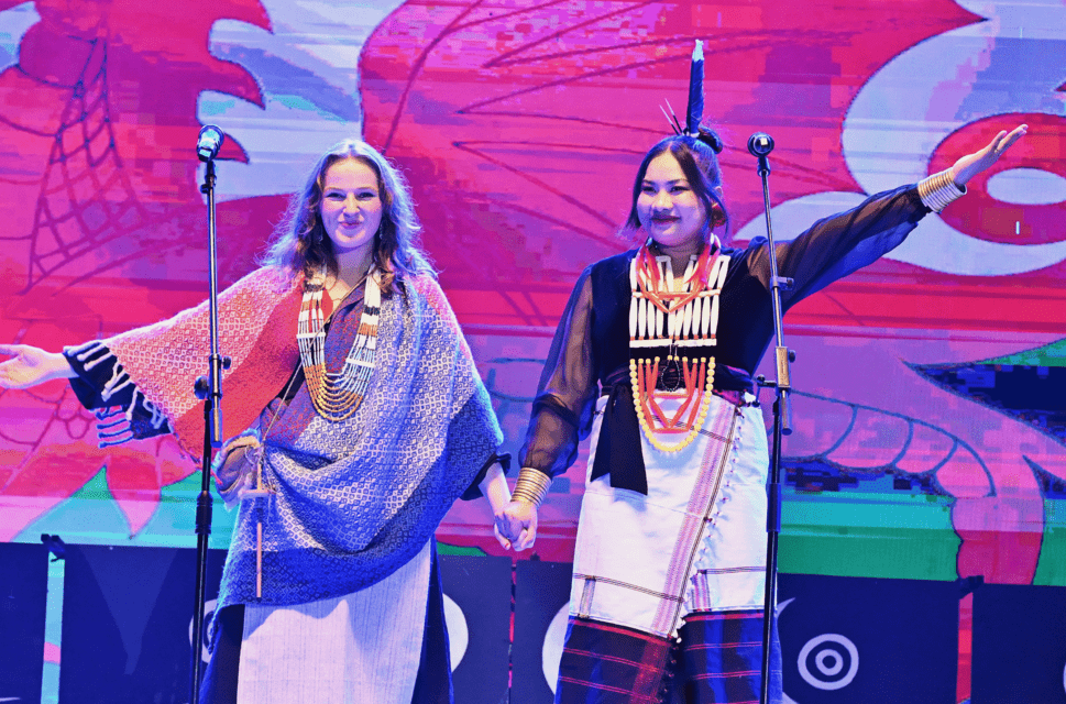 Two women stand on stage looking out to an audience out of shot. They are holding hands and waving to the audience, smiling. They stand against a large Welsh flag and are wearing traditional Indian clothing.