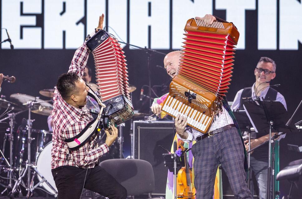 Two musicians, one Scottish and one Mexican, play the accordian energetically on a stage with the rest of the band looking on