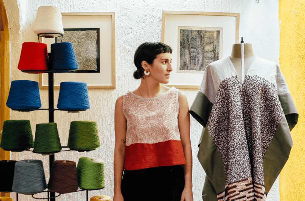 A woman stands between a manequin and a stand holding spools of coloured yarn. She looks to her left, beyond the manequin. She's wearing a gold earrings and a black, white and red striped top, her short dark hair is tied back. There are two framed pictures out of focus hanging on the wall behind her.