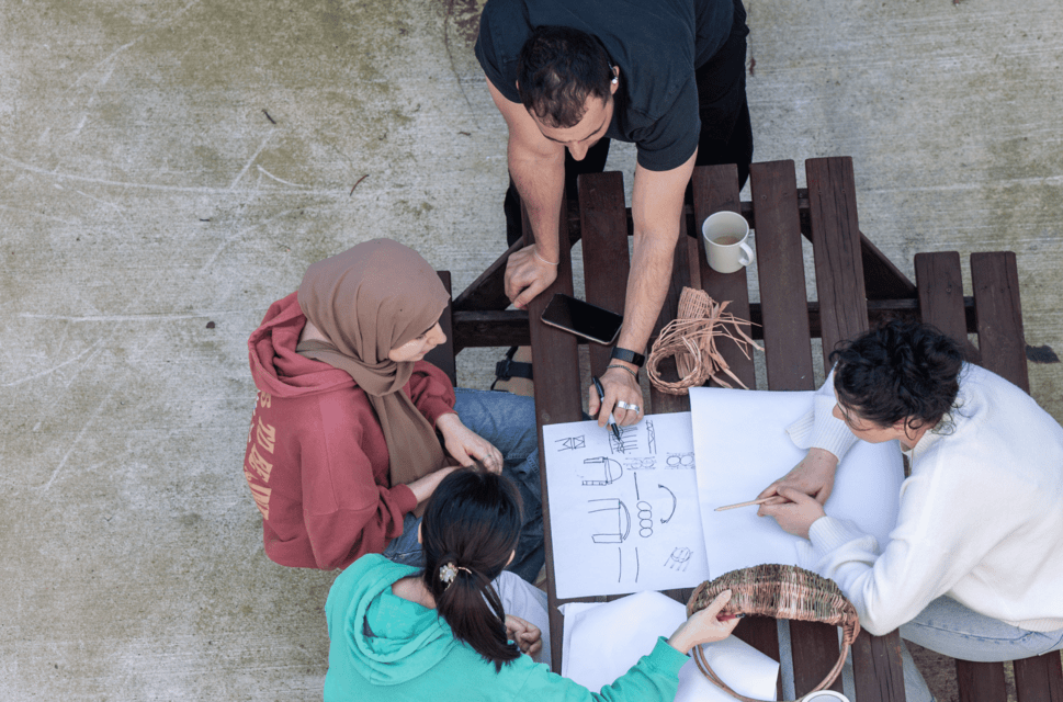 An overhead shot of a group of four people sitting and standing around a wooden picnic bench, looking at papers and crafting materials spread on its surface, seemingly in conversation and working on something together.