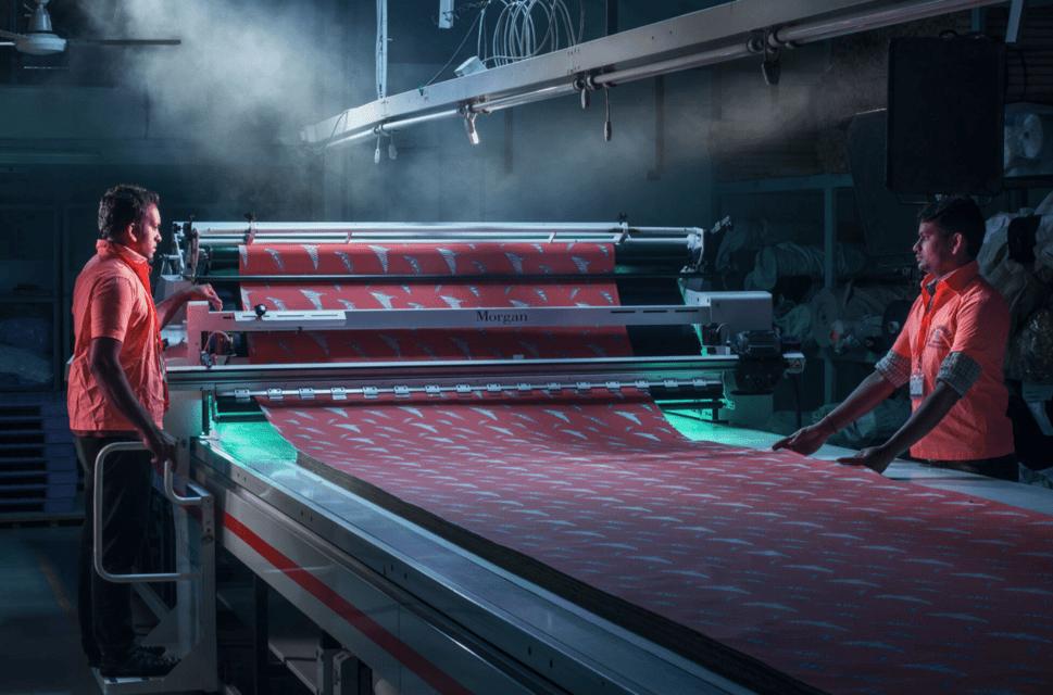 Two men stand either side of a fabric production line in a factory.
