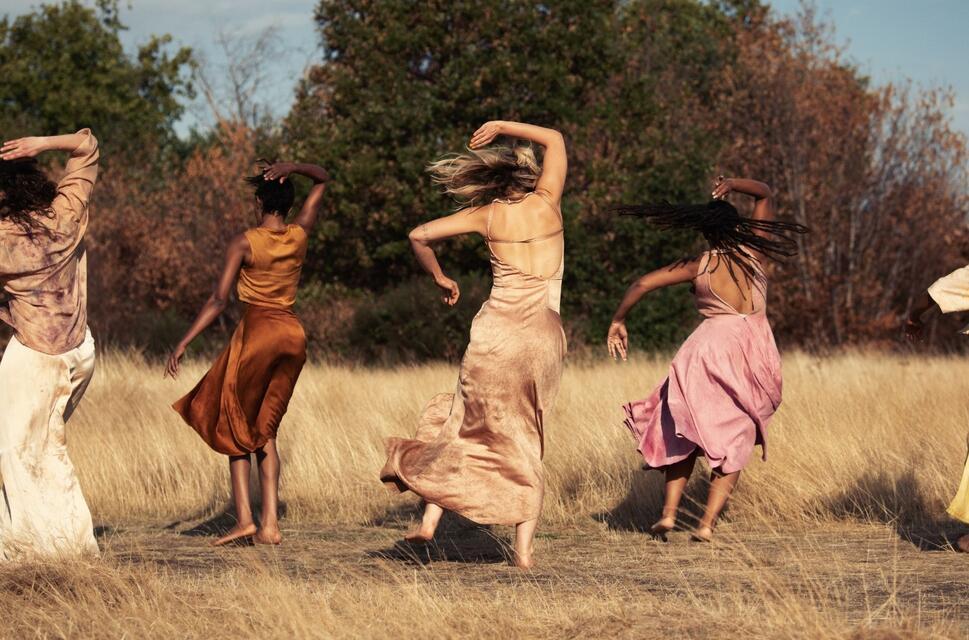 Five women in flowing dresses dance in a field