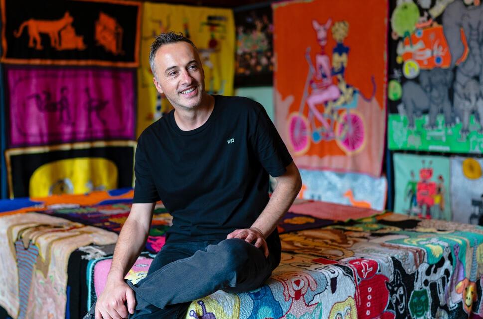 Man smiling, sat on fabric artwork with artwork around him.