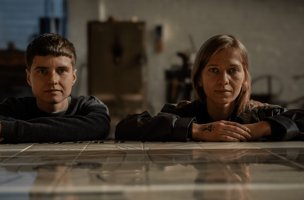 Two people sit alongside each other with their arms resting on a shiny tabletop. They are looking straight ahead, into the camera.