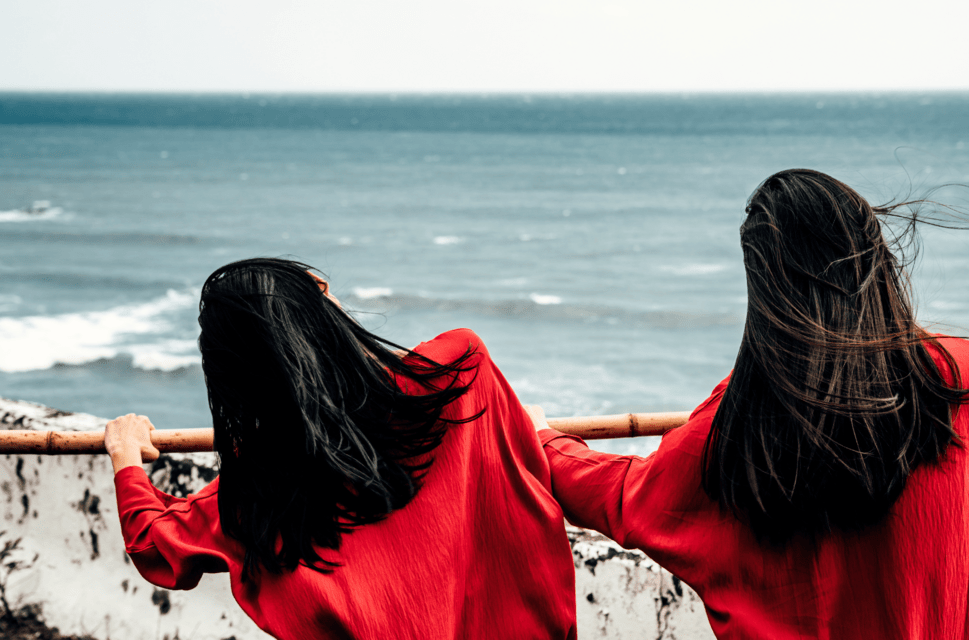 Two girls with long dark hair wearing red stand in the foreground by a seafront, looking out to see holding onto a rusty rail.