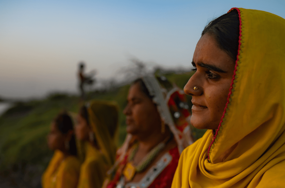 A line of four women stand in a line reaching diagonally left from the right hand side of the image. The woman closest to the camera is in focus and the rest are out of focus, as is the background, which appears to be a rural setting next to water. The women are wearing yellow head coverings.