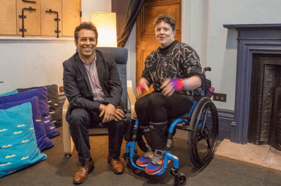 Artistic Director & CEO of Battersea Arts Centre, Tarek Iskander with Jess Thom (Touretteshero) in the new permanent rest space at BAC. They sit alongside each other with a fireplace to their left, and large colourful cushions are piled to their right. Tarek Iskander is seated on a chair, wears a grey suit and brown shoes, and is smiling. Jess Thom is seated in her wheelchair and is wearing a sweatshirt with a graphic print and colourful trainers.