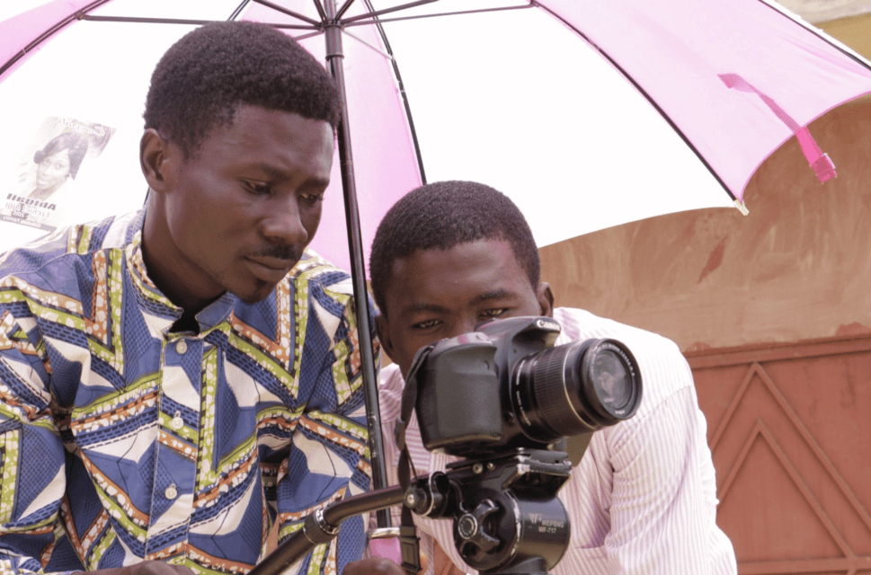 Two men hold a video camera under an umbrella. They are filming something out of frame.