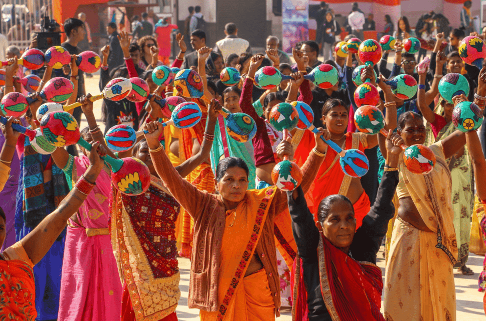 WOW Nepal, 2019. A crowd of women wearing brightly coloured saris stand with their arms raised holding what looks like large papier mache maracas.