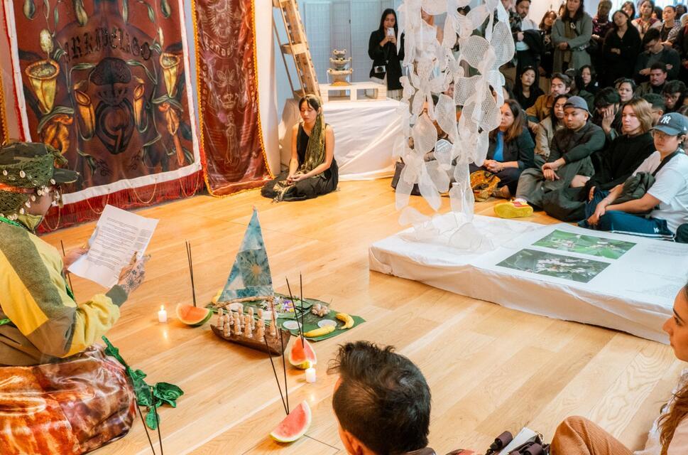 A performer reads to an audience, seated on the floor