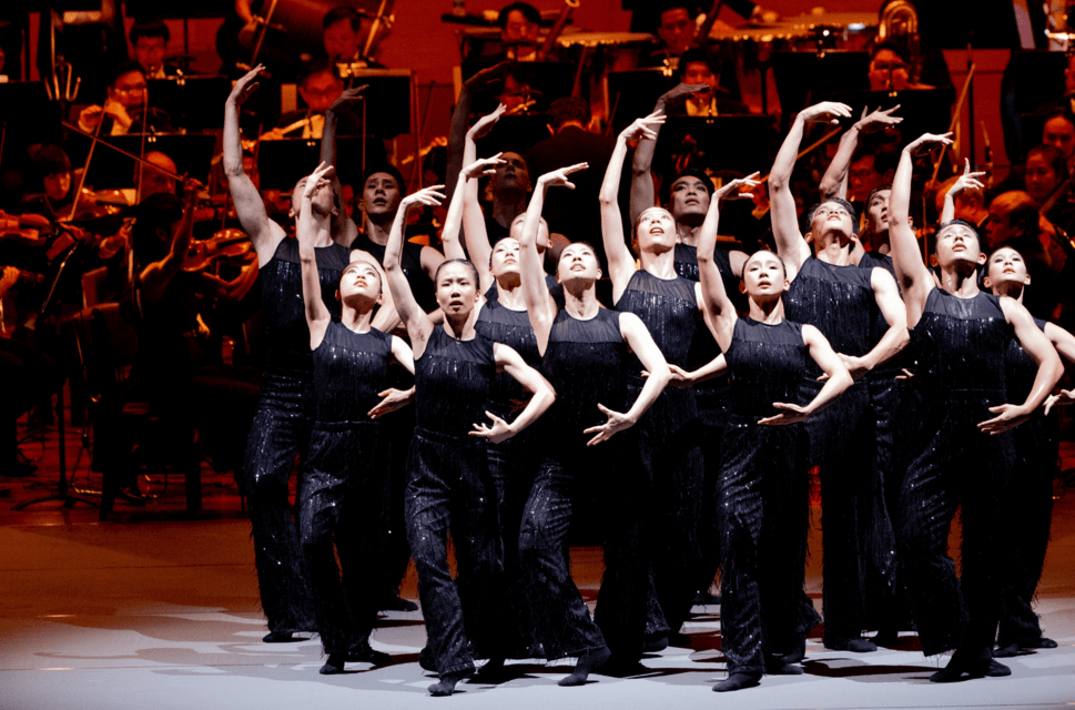 A group of dancers dressed in black stand in the same balletic pose on stage, with their right arms held above their heads, and their left arms below, looking upwards 