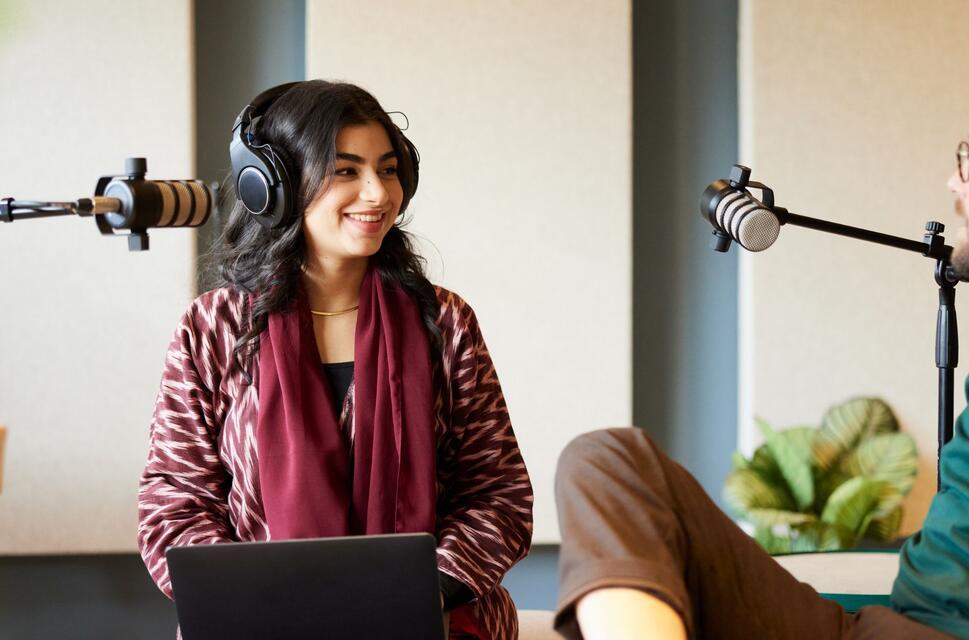 A woman and man with headphones speak into mics for a podcast recording