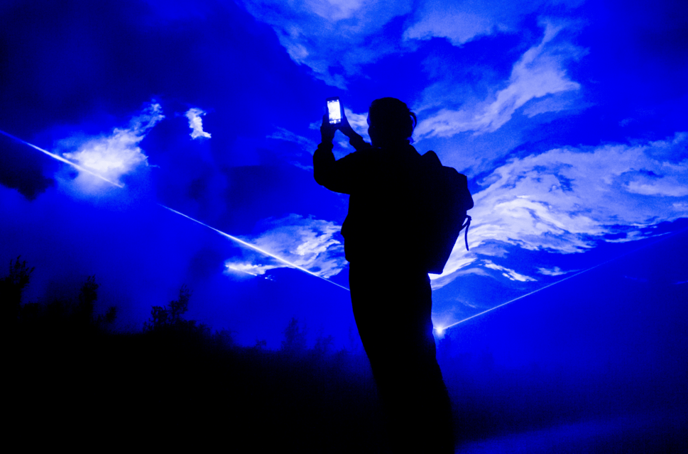 A figure stands silhouetted against a projection of bright blue and white swirling clouds. The figure holds up a mobile phone with a lit-up screen, as if taking a photo.
