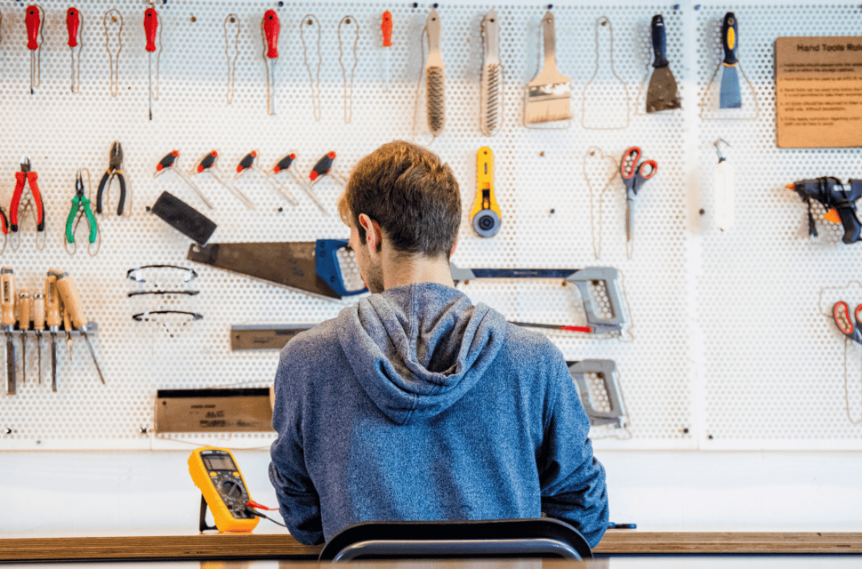 A man sits at a table or workbench with his back to the camera. In front of him is a wall with various tools hanging on it.